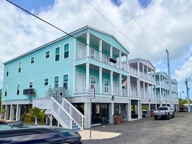 Pastel turquoise beachfront house on stilts with white porches and stairs, parked cars below.