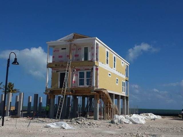 Elevated yellow beach house under construction on stilts beside the ocean, with sand and blue sky.