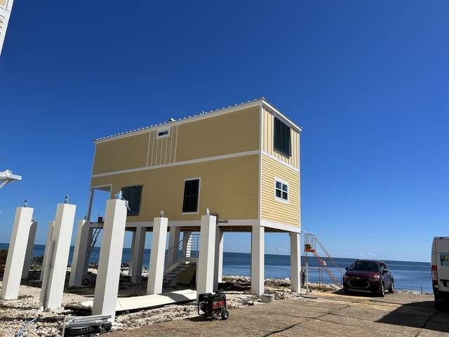 Elevated yellow house on stilts under construction by the beach, with cars and white pilings nearby.