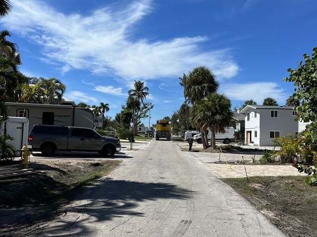Sunny residential street with palm trees, parked vehicles, and white houses under a blue sky.