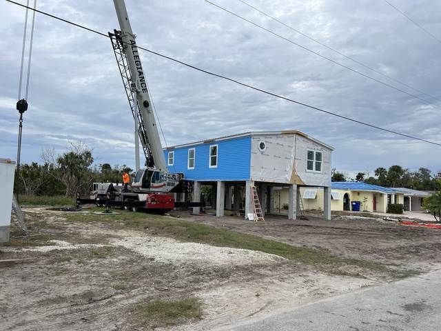 Crane lifting a blue house section at a construction site under a cloudy sky.