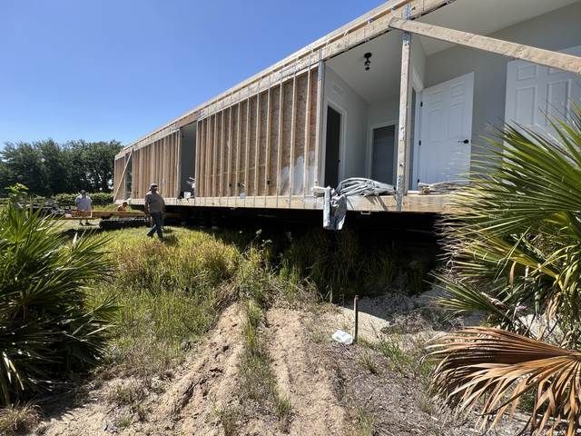 Partially built house on stilts over a grassy, sandy area with one person walking nearby.