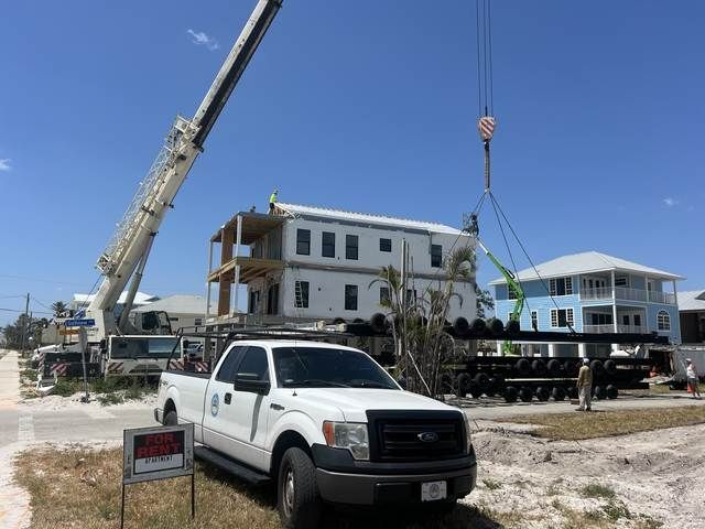 Construction site with white pickup truck, crane, and a raised white house under a blue sky.