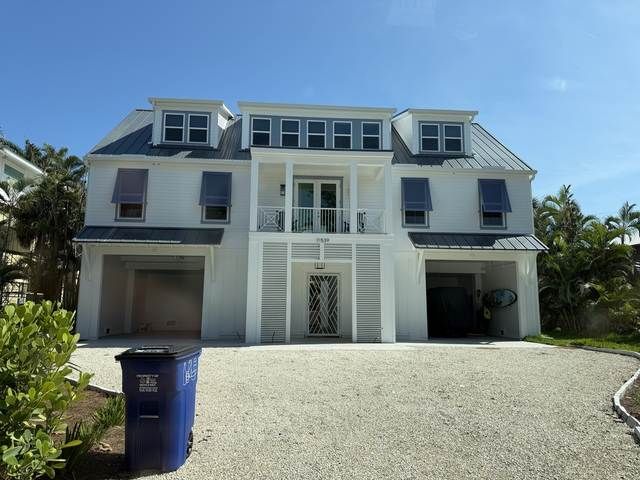 Large white two-story house with a gravel driveway and blue trash bin under a clear sky.