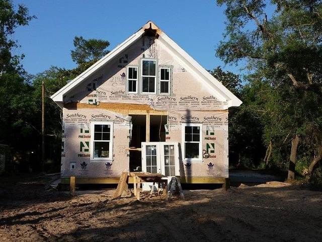 Two-story house under construction with exposed framing, white wrap, and a dirt yard in front.