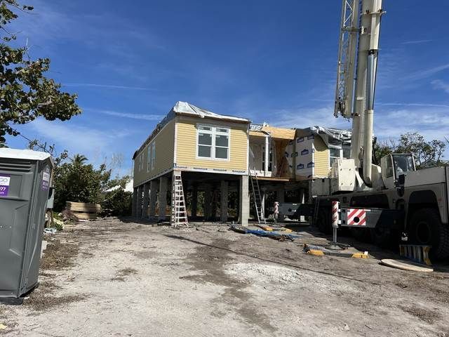 Yellow house on stilts under construction beside a utility truck on a dirt lot.