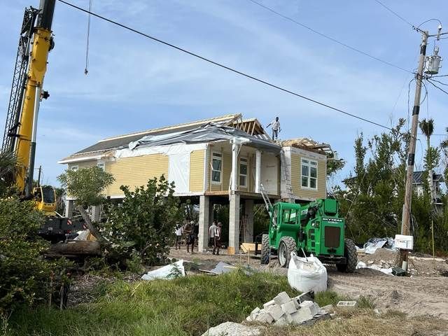 House under construction with yellow siding, roof framing exposed, and heavy equipment on-site.