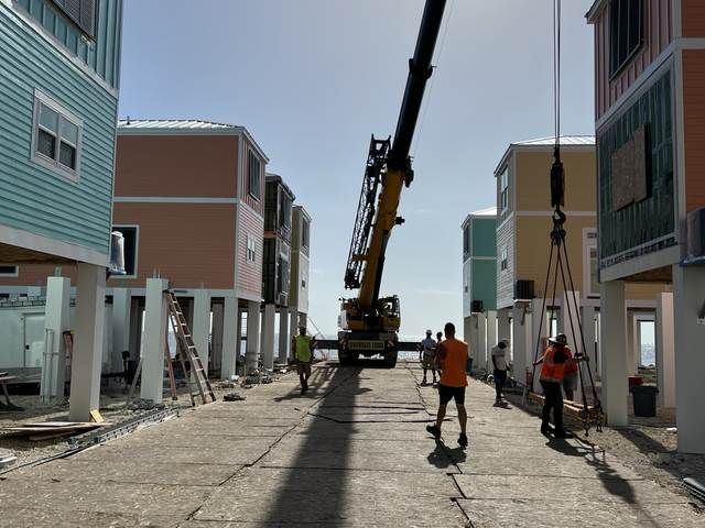 Construction workers guiding a crane in a narrow street between modern houses, with utility poles and rubble nearby.