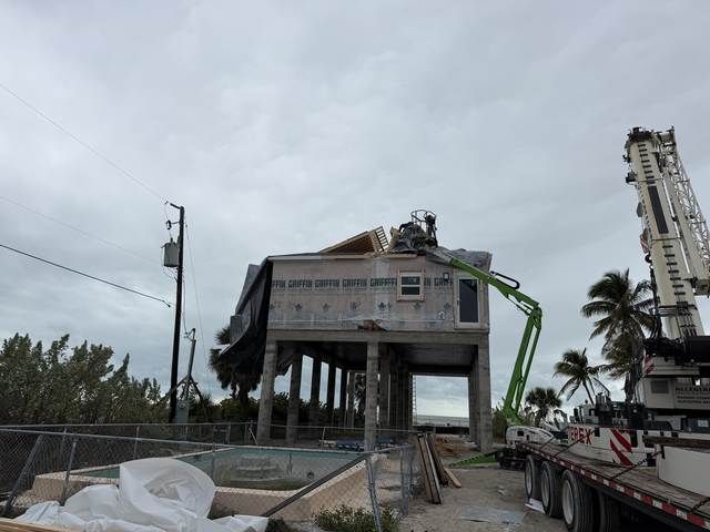 Damaged house on stilts being lifted by crane after a storm, with debris and cloudy sky around it.
