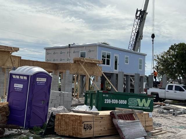 Portable toilet beside a construction site with a raised blue house, crane, and building materials.