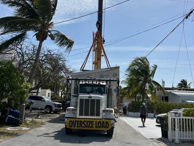 Oversized load truck carrying a tall utility pole on a residential street with palm trees and a person walking nearby.