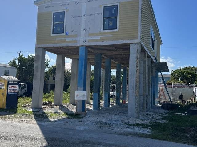 Elevated house under construction on concrete stilts, with exposed framing and nearby portable toilet.