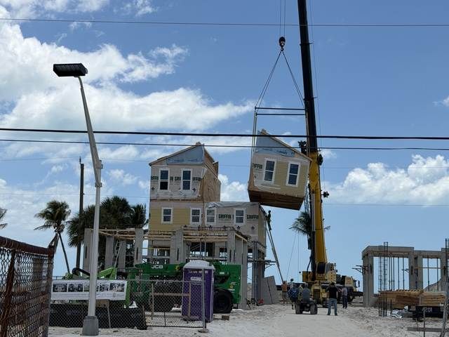 Storm-damaged beach houses lifted on stilts, with a leaning utility pole and construction equipment nearby.