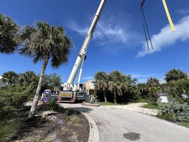 Crane truck lifting a long boom over a landscaped road with palm trees and blue sky.