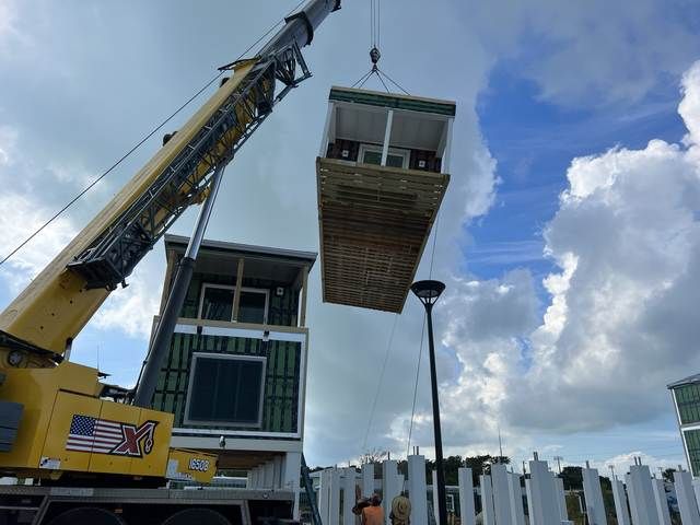Crane lifting a modular building section against a blue sky near a waterfront.