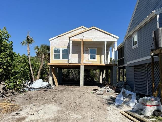 Raised house under construction on stilts with a front porch and dirt driveway, surrounded by debris.