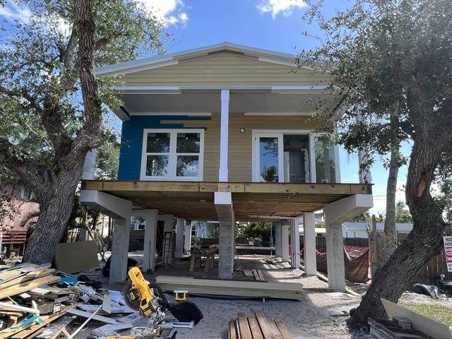 Raised house under construction, yellow siding, supported on stilts amid debris and trees.