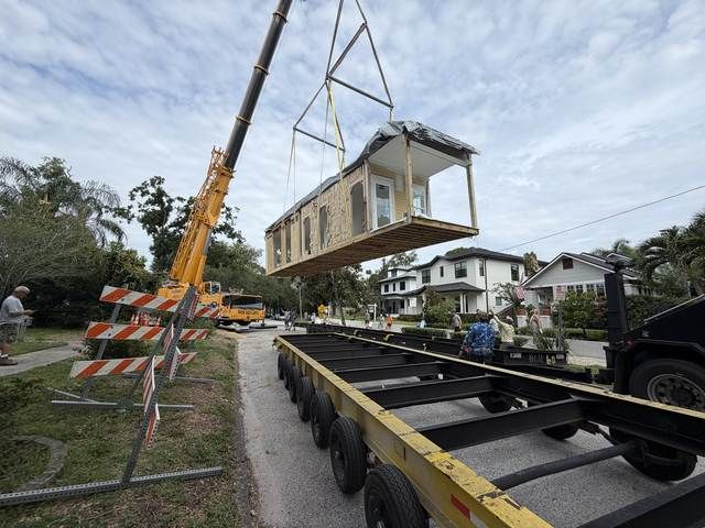 Crane lifting a modular home onto a flatbed trailer on a residential street.