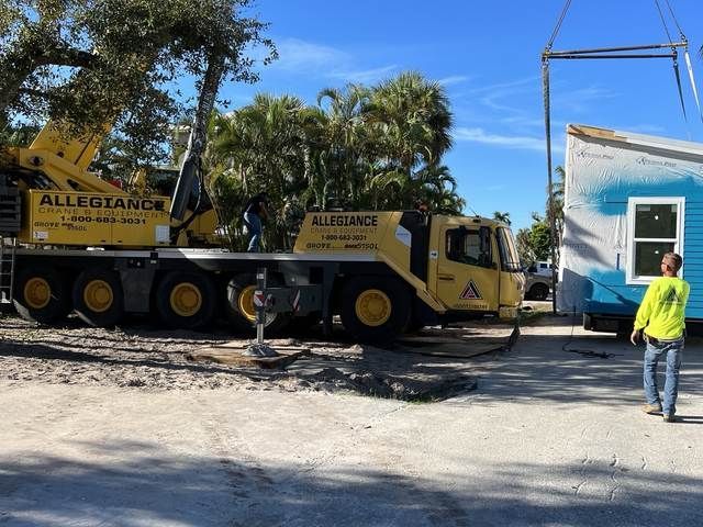 Yellow Allegiance crane truck beside a white building, with a worker in a neon shirt nearby.