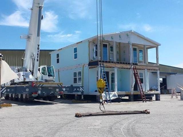 Portable home being installed with crane on construction site, with workers and ladders near a two-story house.