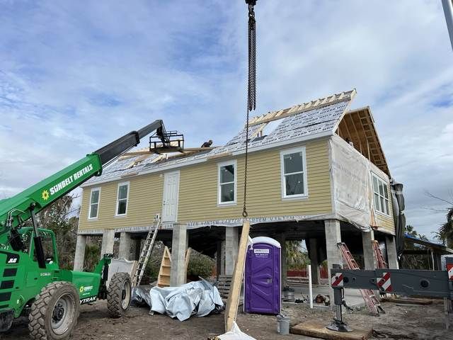 House raised on stilts at a construction site with a green crane and work equipment nearby.