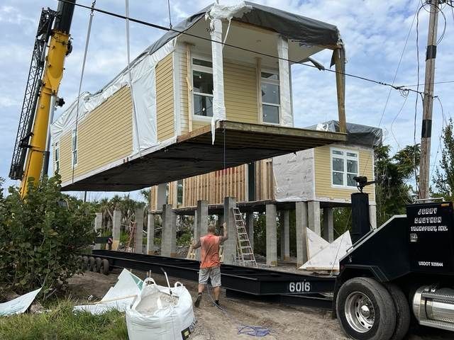 House being lifted by crane during relocation, with workers and a truck at a roadside site.