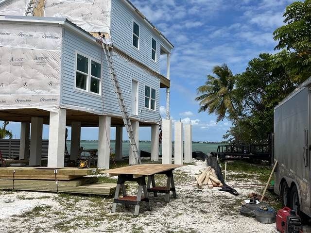 Stilt house under construction beside a trailer, with palm trees and blue sky in the background.