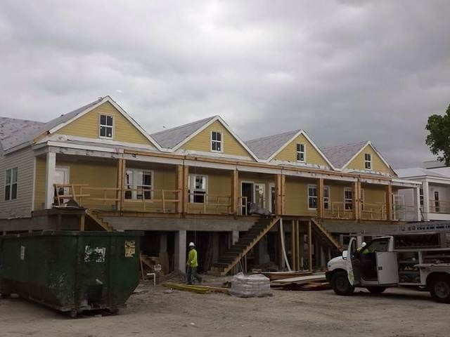 Elevated yellow house under construction with workers, dumpster, and utility truck on a cloudy day.