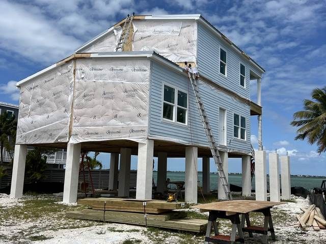 Elevated coastal house under construction on stilts with exposed insulation and blue sky.