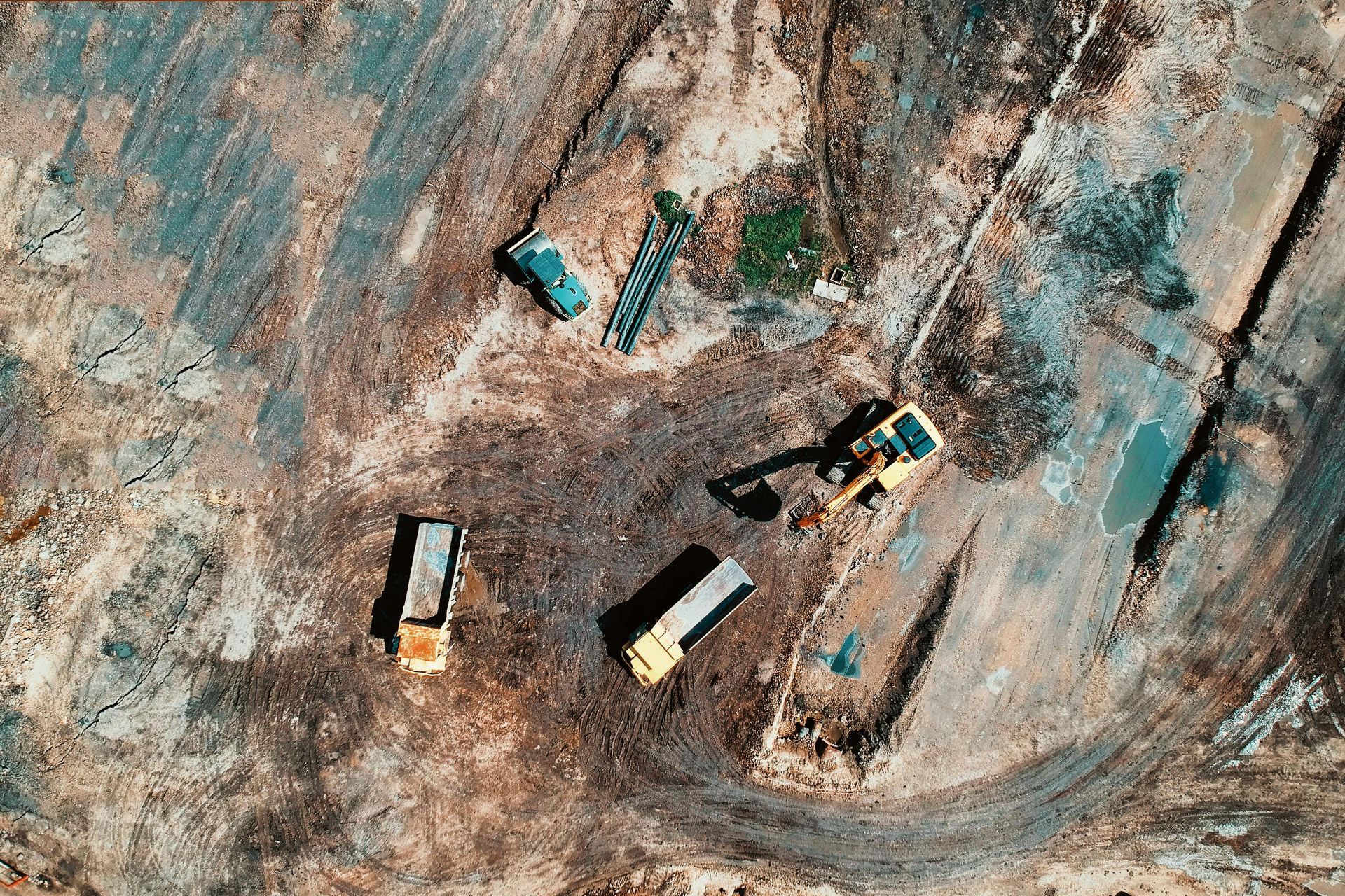 Aerial view of construction site with trucks, excavator, and various earth tones.
