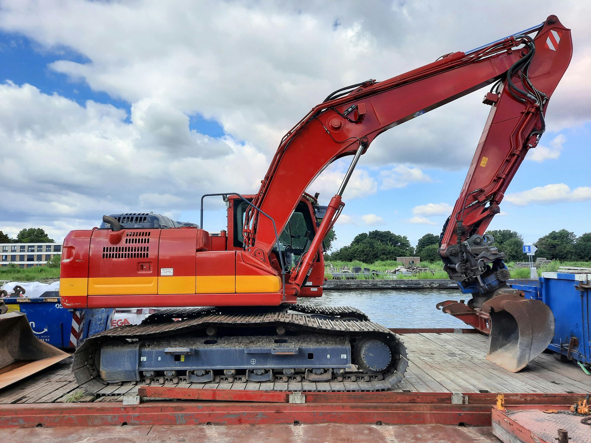 Red excavator on a barge, operating near a body of water, under a cloudy sky.