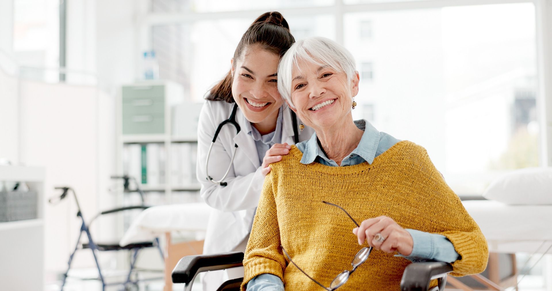 Doctor with stethoscope smiles behind an elderly woman in a wheelchair, both smiling; bright room.