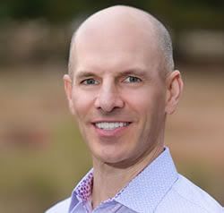 Bald man smiling, wearing a light blue collared shirt.