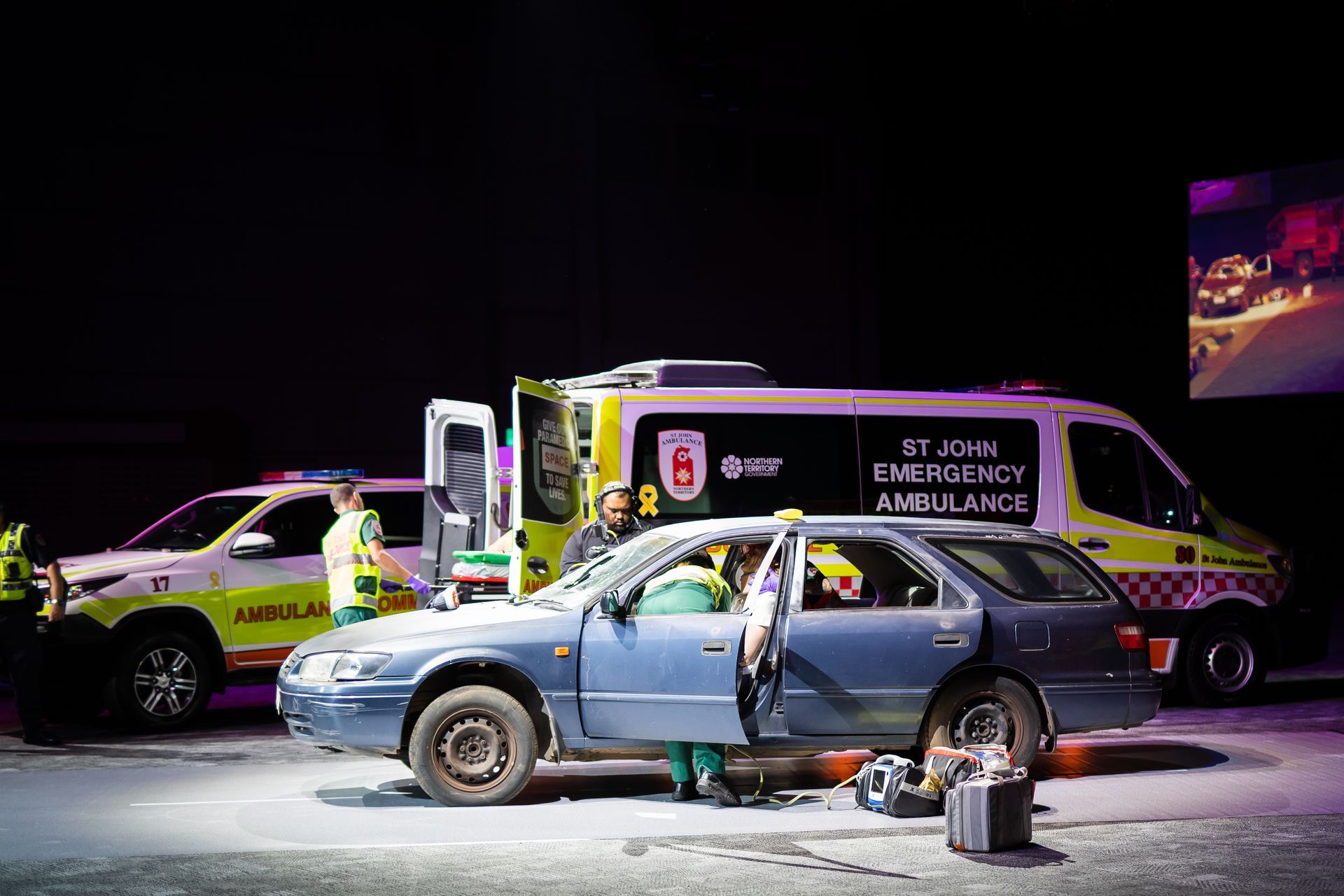 A blue car is parked next to an ambulance on a stage.
