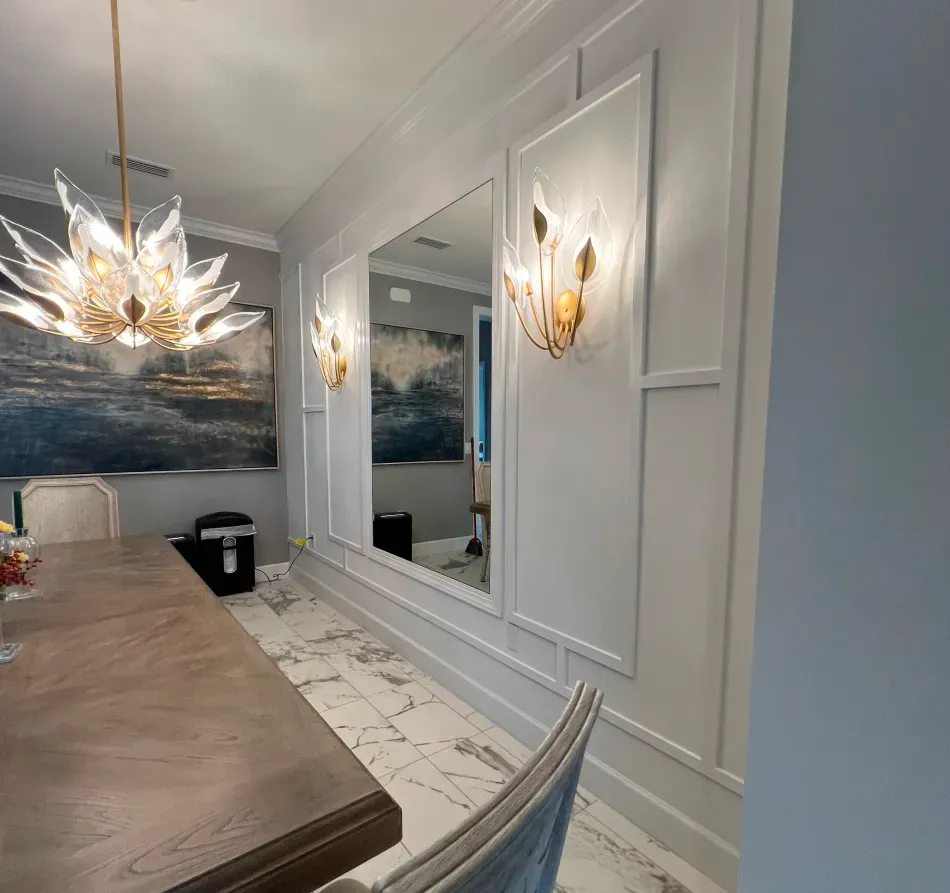 Dining room with white paneled walls, large mirror, gold sconces, and a floral chandelier.