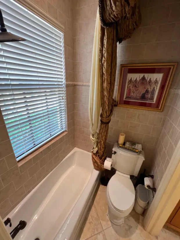 Bathroom with bathtub, toilet, window with blinds, and decorative curtain. Beige tile.