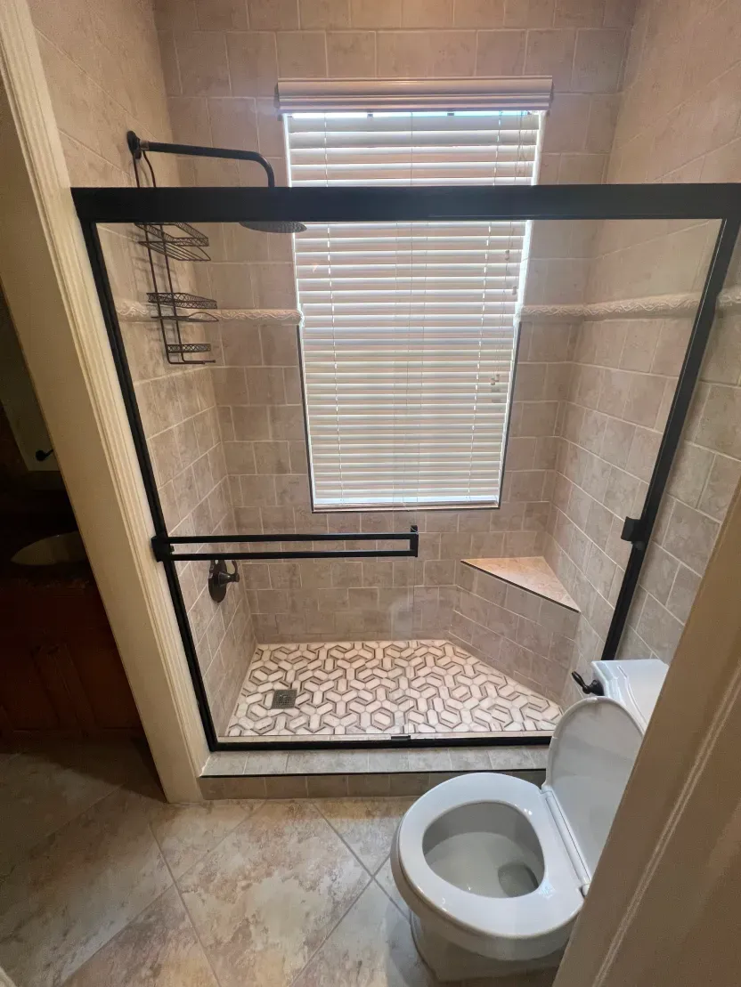 Bathroom with glass-door shower, toilet, and window with blinds. Tile walls and floor. Black shower fixtures.
