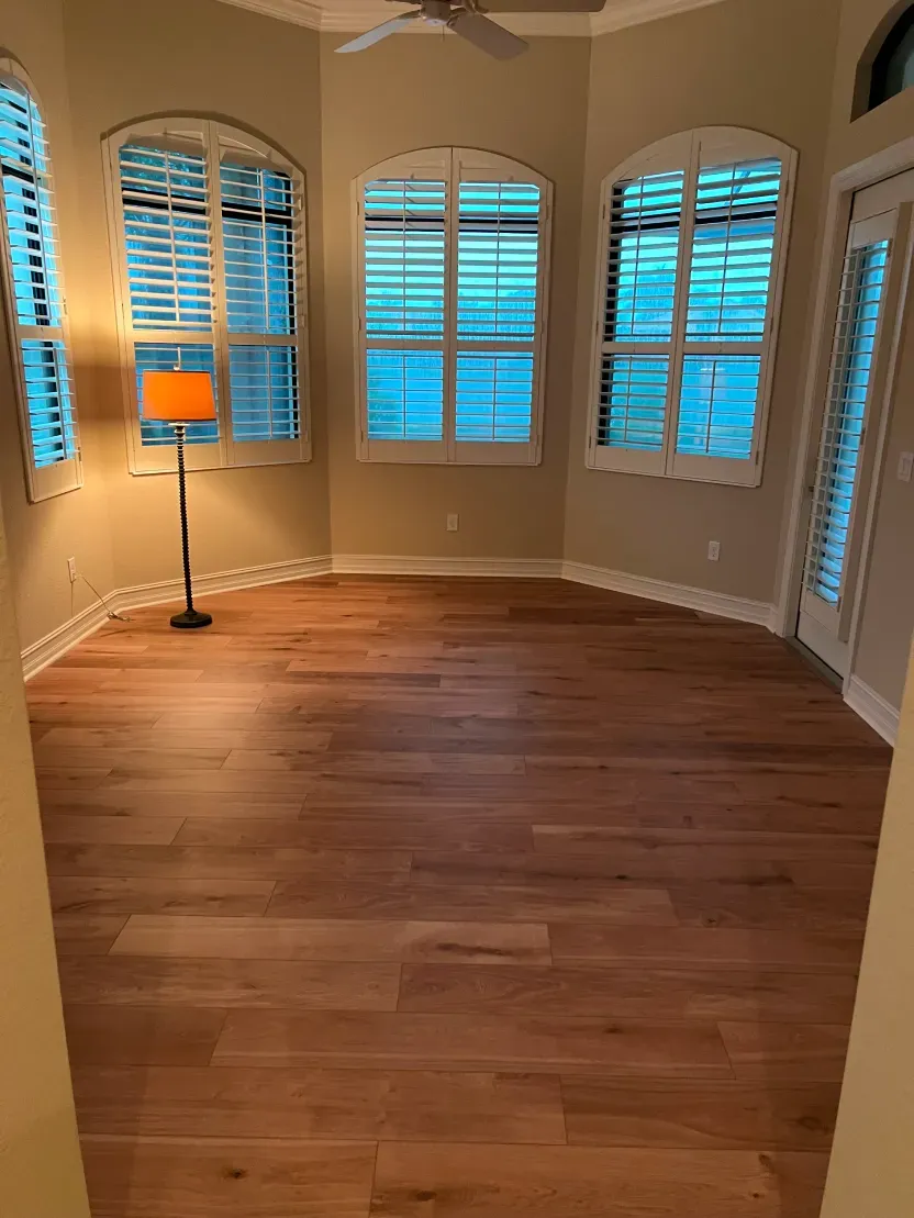 Empty room with hardwood floors, three arched windows with shutters, and a floor lamp.