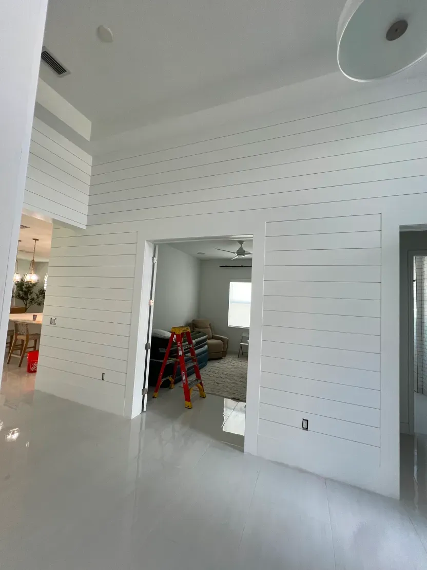 Interior with white shiplap walls, doorway, and glossy floors; a room with a red ladder is visible.