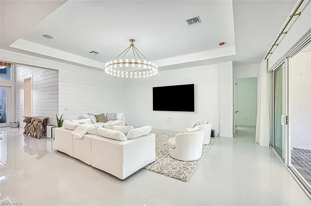 A modern, all-white living room featuring a large sofa, two armchairs on a patterned rug, a wall-mounted TV, and chandelier.