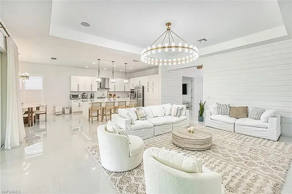 Bright, open-concept living room and kitchen featuring white furniture, a large round chandelier, and a shiplap wall.