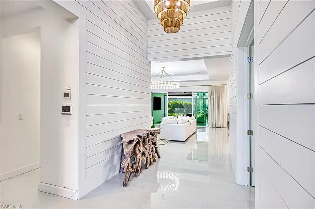 A white, minimalist foyer features horizontal shiplap walls, a sculptural wooden console table, and a tiered chandelier.