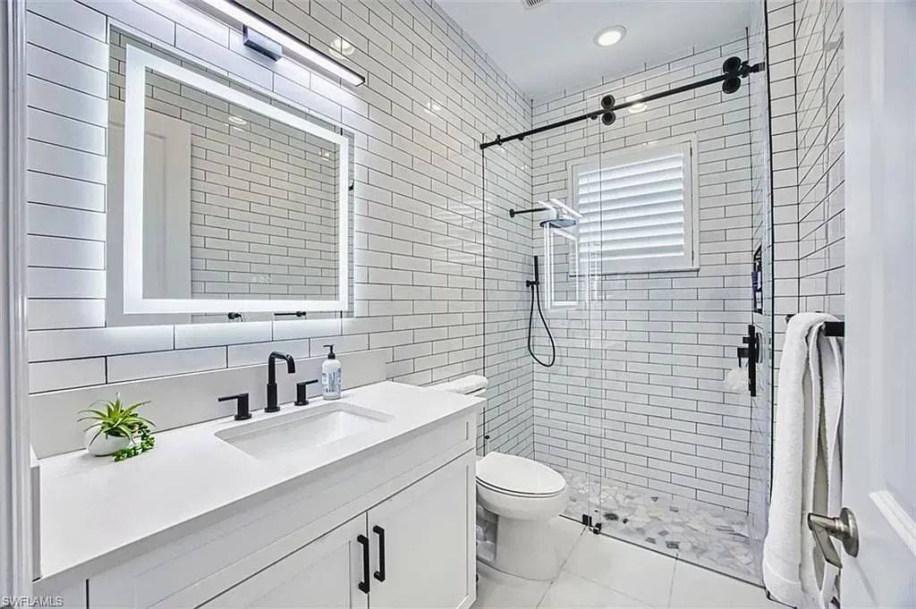A modern bathroom features a white vanity, a large lighted mirror, and a glass-enclosed shower with decorative wall tiles.