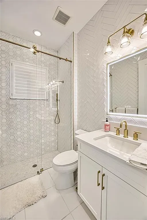 A bright, modern bathroom featuring a white vanity with gold hardware, a glass-enclosed shower, and a toilet.