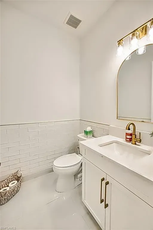 A bright, modern bathroom with a white vanity, gold faucet, arched mirror, tiled walls, and a toilet next to a basket.