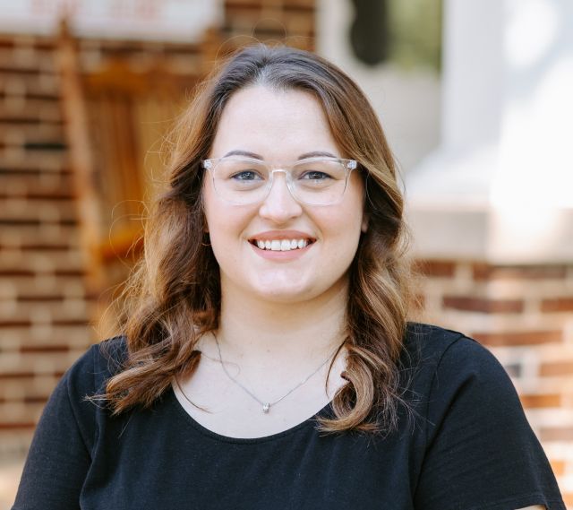 A woman wearing glasses and a black shirt is smiling in front of a brick building.