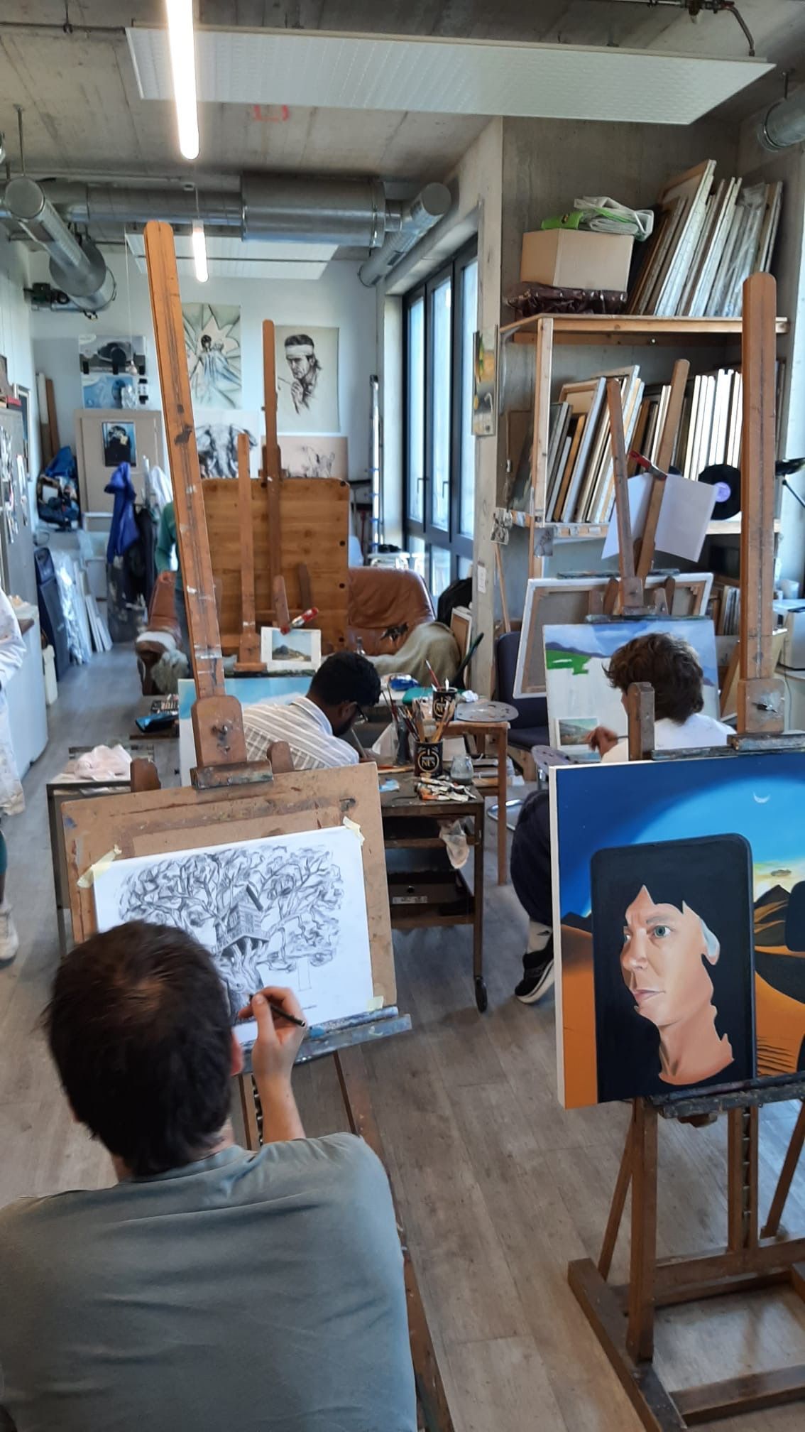 Atelier d'art avec plusieurs chevalets. Des artistes dessinent ou peignent devant les chevalets. Des étagères en bois accueillent les fournitures en arrière-plan.