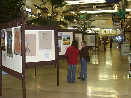Exposition d'art dans un couloir lumineux d'un centre commercial. Deux personnes contemplent des œuvres encadrées sur des panneaux d'exposition.