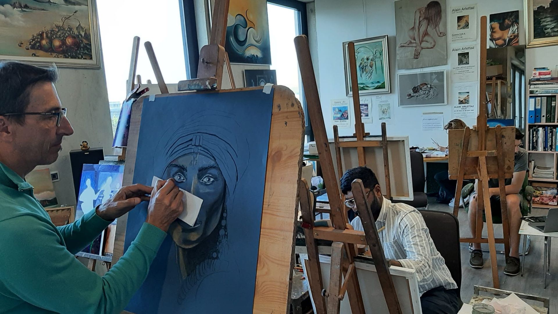 Un homme peint un portrait sur une toile bleue dans un atelier. Une autre personne dessine sur un chevalet. Œuvres d'art et étagères en arrière-plan.
