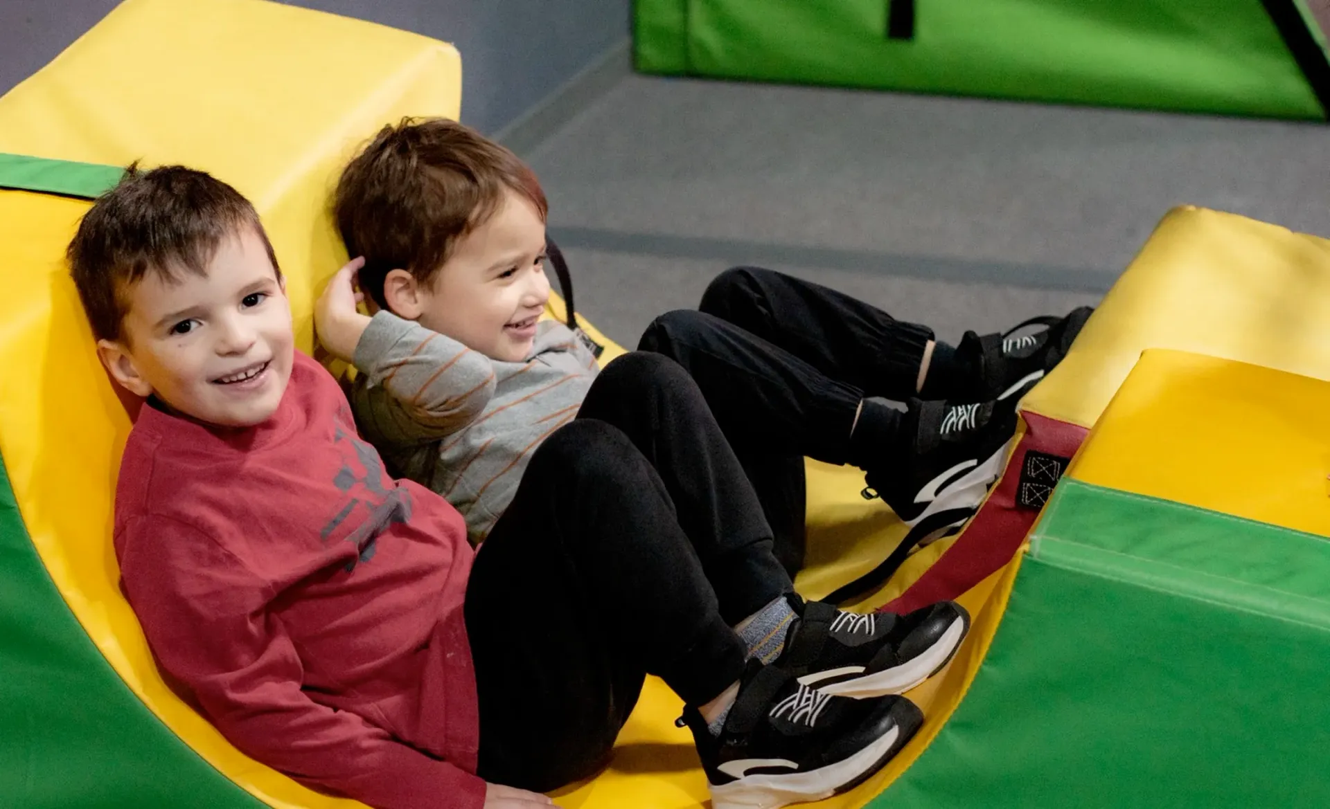 Two children sitting on colorful play blocks, smiling in a play area